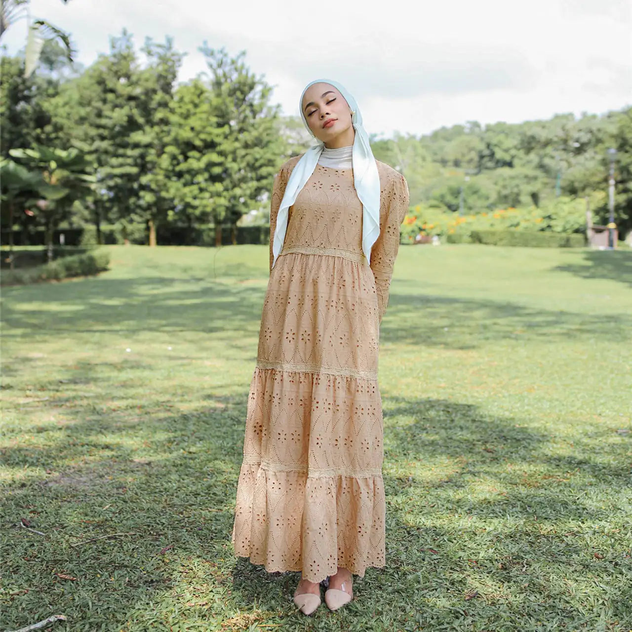 Model weraing a white scarf and beige dess with trees and green lawn in the background