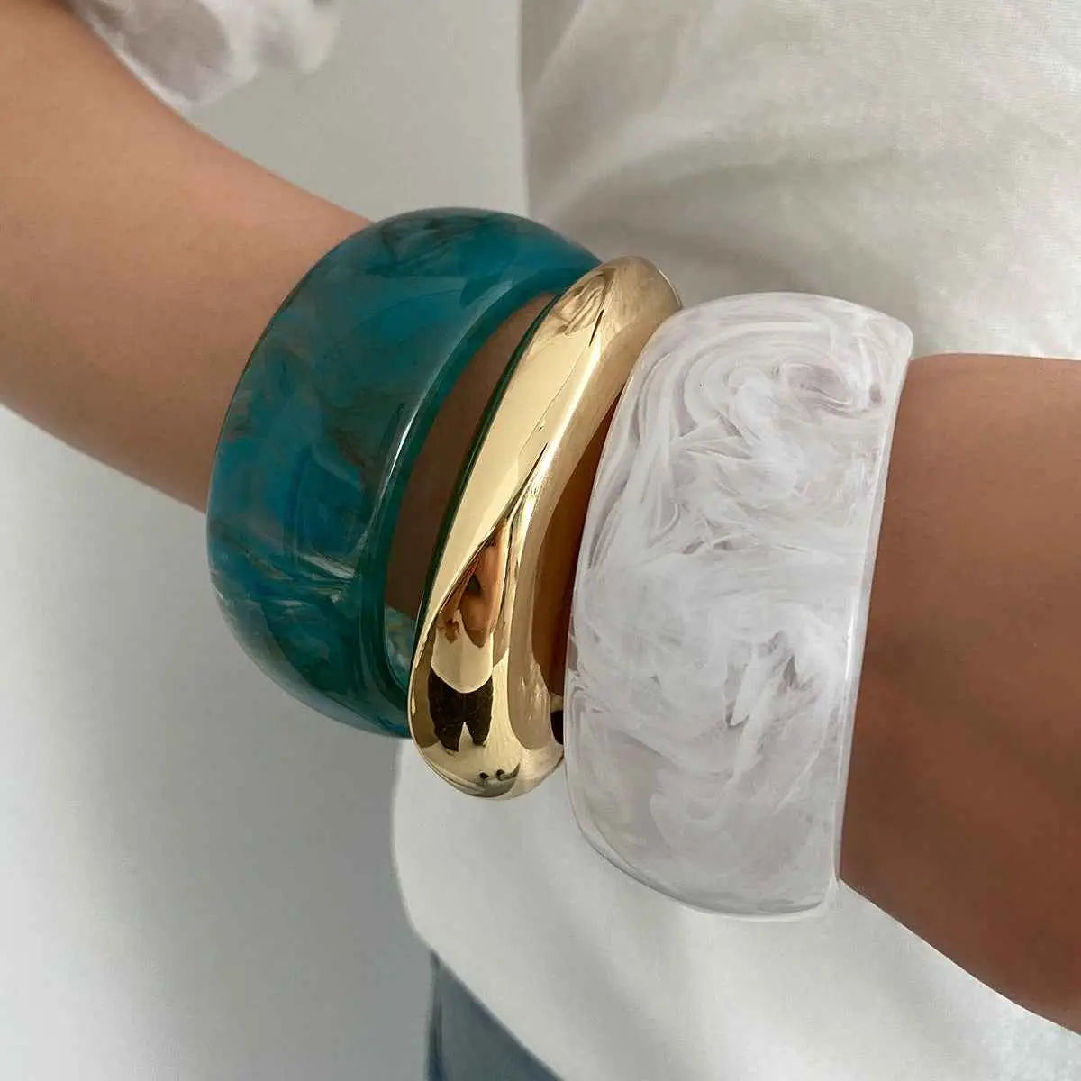 Three stacked bangles on wrist featuring a marbled translucent resin white bangle teal translucent and polished gold designs