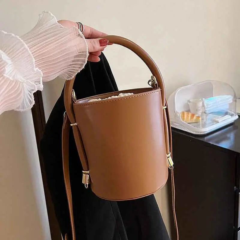 Structured tan leather cylinder bucket bag with drawstring inner, curved top handle, adjustable strap, and gold-tone hardware